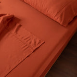 An orange bed with Linenly's Bamboo Sheet Set in Copper Sunset, luxuriously smooth and neatly arranged, features two pillows. The blanket's ultra-soft fabric and detailed stitching are partially revealed, creating an inviting scene on a wooden floor.
