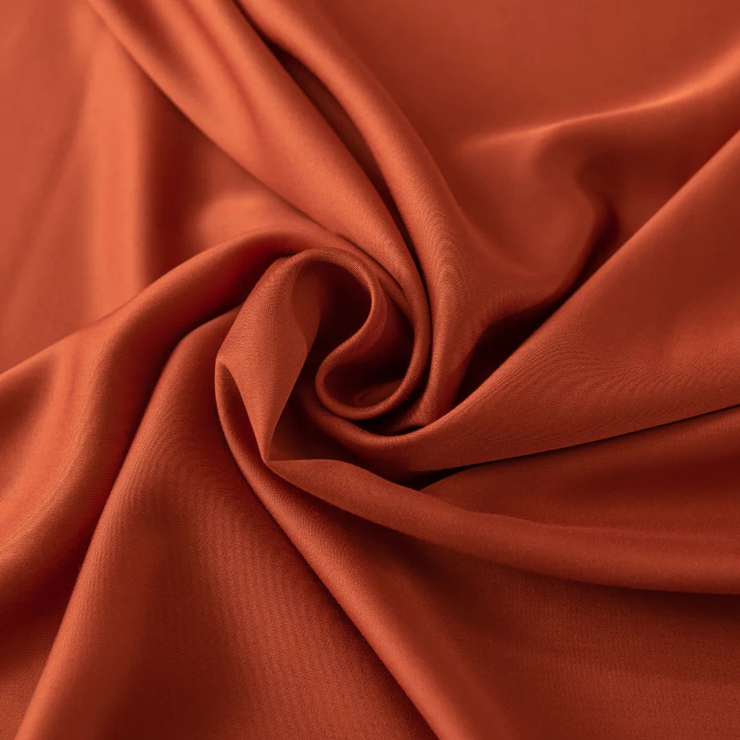 The Bamboo Sheet Set - Copper Sunset from Linenly features rust-hued, silky-smooth bamboo fabric elegantly folded into a swirling pattern at the center.