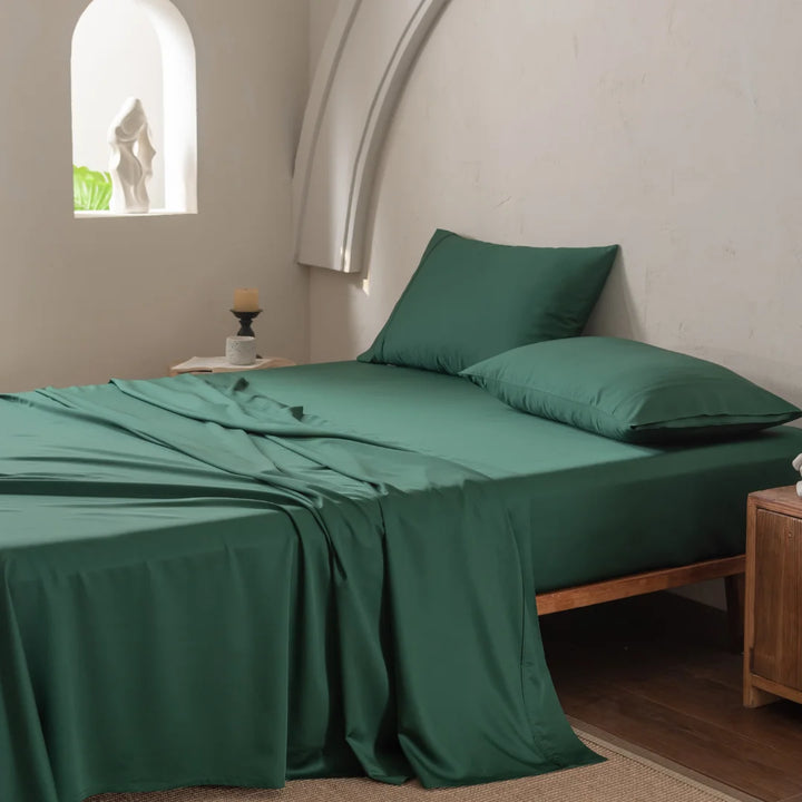 A neatly made bed with Linenly's ultra-soft Bamboo Sheet Set in Evergreen boasts a silky-smooth texture. Light walls and a small wooden nightstand complement an arched niche with a sculpture, while soft lighting enhances the serene atmosphere of this luxurious retreat.