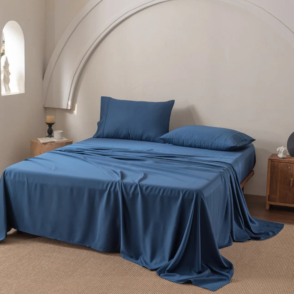 A neatly made bed with Linenly's ultra-soft Bamboo Sheet Set in Midnight and two pillows, set against a cream wall with an arched detail. Wooden nightstands hold a candle and decor items. A beige carpet covers the floor, complementing the sheets perfectly.