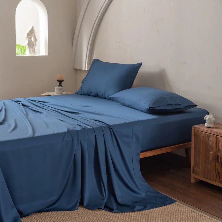 A neatly made bed showcases the ultra-soft Linenly Bamboo Sheet Set - Midnight, complemented by two pillows against a light-colored wall. A wooden nightstand displays a small sculpture and candle, while soft natural lighting streams in from a nearby window.