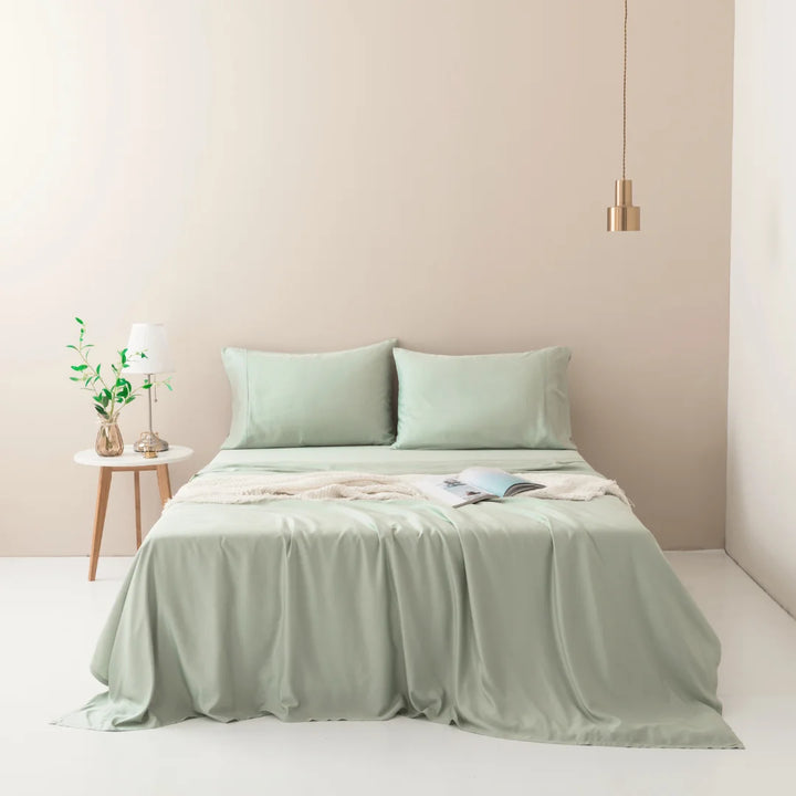 A serene bedroom features a neatly made bed with Linenly's soft sage bamboo sheet set, crafted from sustainable bamboo fibers and luxuriously soft to the touch. Above, a hanging gold pendant light illuminates the space.