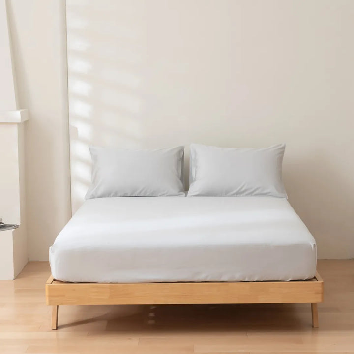 A minimalist bedroom with a neatly made bed, featuring a Luxe Sateen Fitted Sheet in Silver from Linenly and two pillows on a wooden bed frame, illuminated by soft natural light casting shadows on a plain wall.