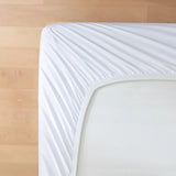 A close-up of the Linenly Luxe Cotton Sateen Fitted Sheet in white, smoothly covering a mattress corner on a light wooden floor.