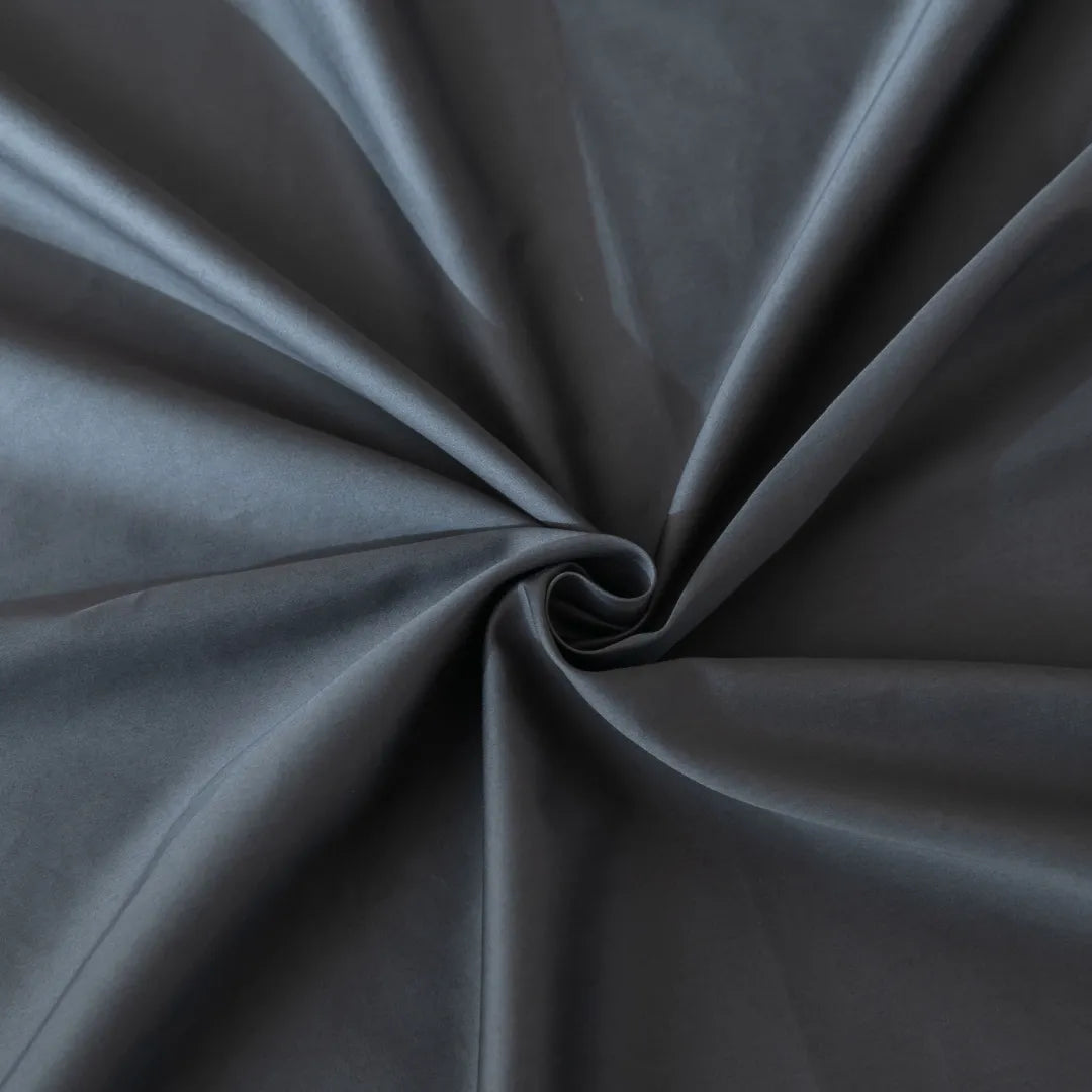 The Luxe Cotton Sateen Flat Sheet - Charcoal from Linenly features smooth, dark gray fabric twisted into a spiral with soft folds. Crafted from 100% cotton sateen, it offers a silky, glossy texture and elegant sheen.