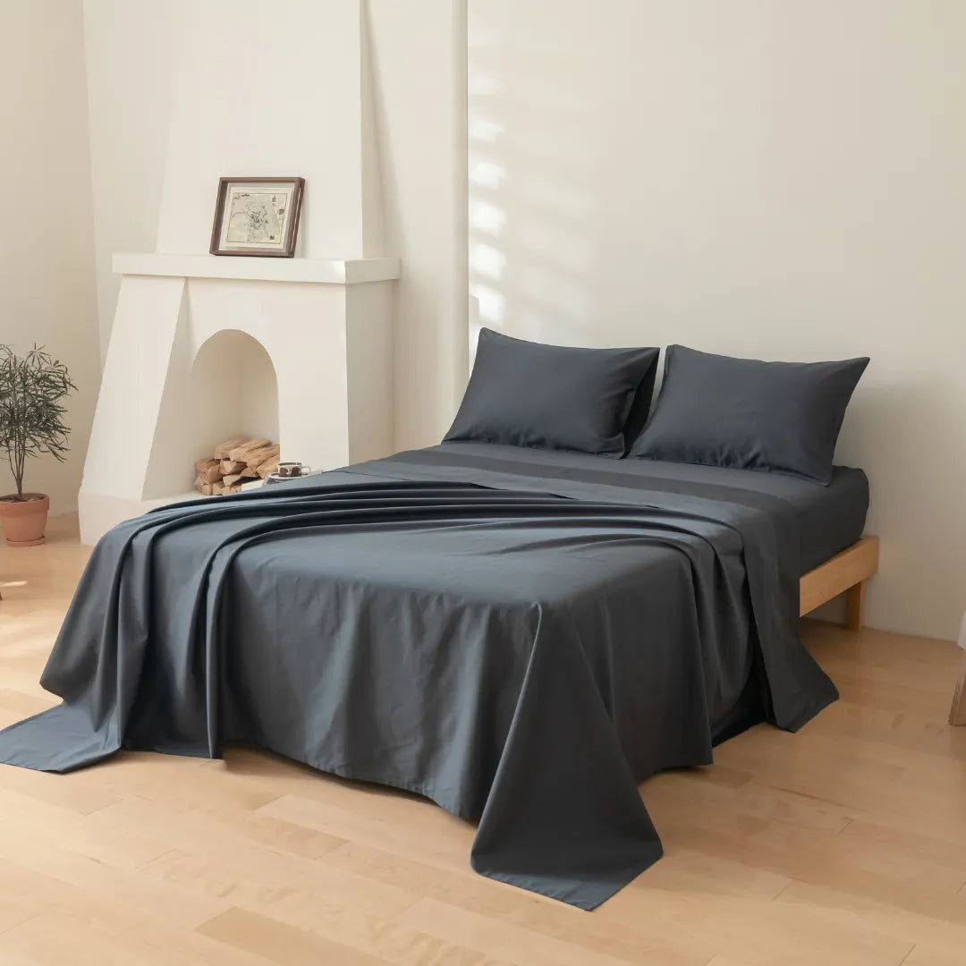 A minimalist bedroom features a neatly made bed with the Linenly Luxe Cotton Sateen Flat Sheet in Charcoal and dark gray pillows. The 100% cotton sateen offers a subtle sheen, set on light wooden floors beside a white fireplace and greenery.