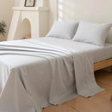 A neatly made bed features the Linenly Luxe Cotton Sateen Flat Sheet in Silver and two pillows on a wooden frame. Light walls, floors, a small plant, and framed picture complete the bright, minimalistic room’s serene atmosphere.