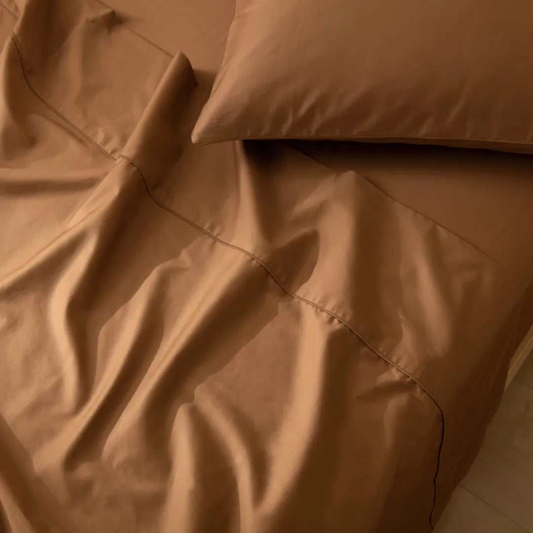 A close-up of a bed features the Linenly Luxe Cotton Sateen Flat Sheet in terracotta and a matching pillow, highlighting the smooth texture and elegant folds of its long staple cotton fabric.