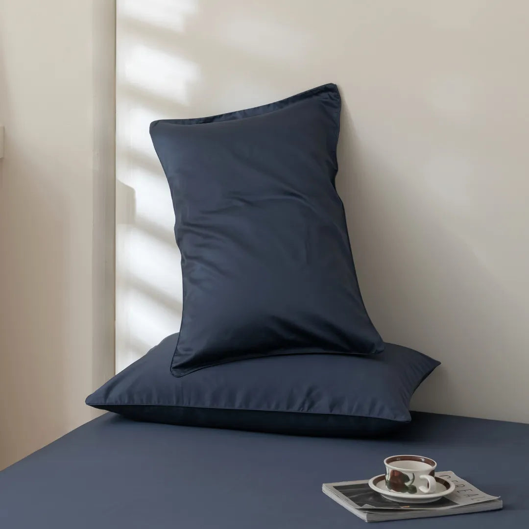 Two Luxe Cotton Sateen Pillowcases in Midnight by Linenly are stacked on a matching bedsheet near a light wall. A cup and saucer rest on a closed book in the foreground, with sunlight casting gentle shadows across the scene.