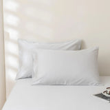 Two Luxe Cotton Sateen Pillowcases in Silver by Linenly are stacked on a neatly made bed with white sheets. Sunlight filters through blinds, casting soft shadows. A book corner and a cup on a saucer rest at the bed’s edge.