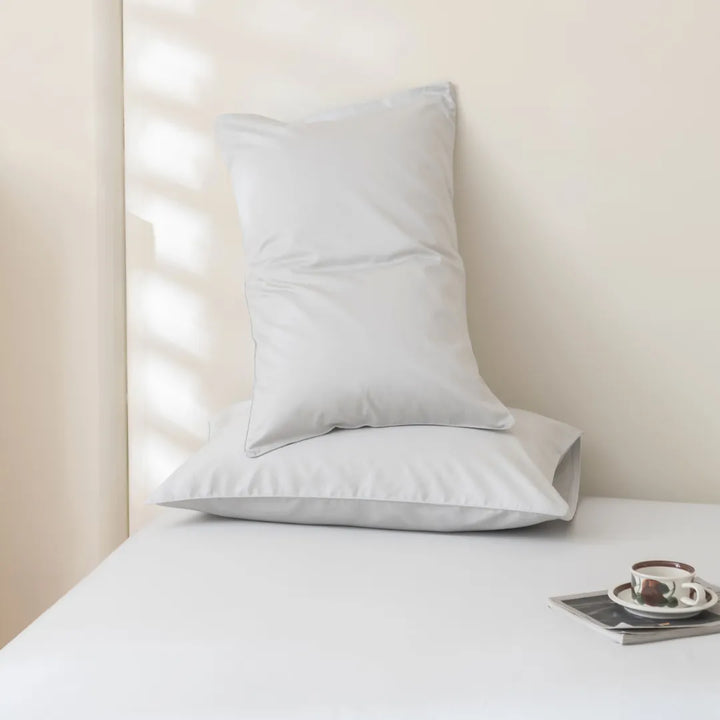 A serene corner with a neatly arranged Luxe Sateen Pillowcase Set - Silver by Linenly on a white surface, accompanied by a magazine and a cup of coffee on a small tray, beside a window with soft light.