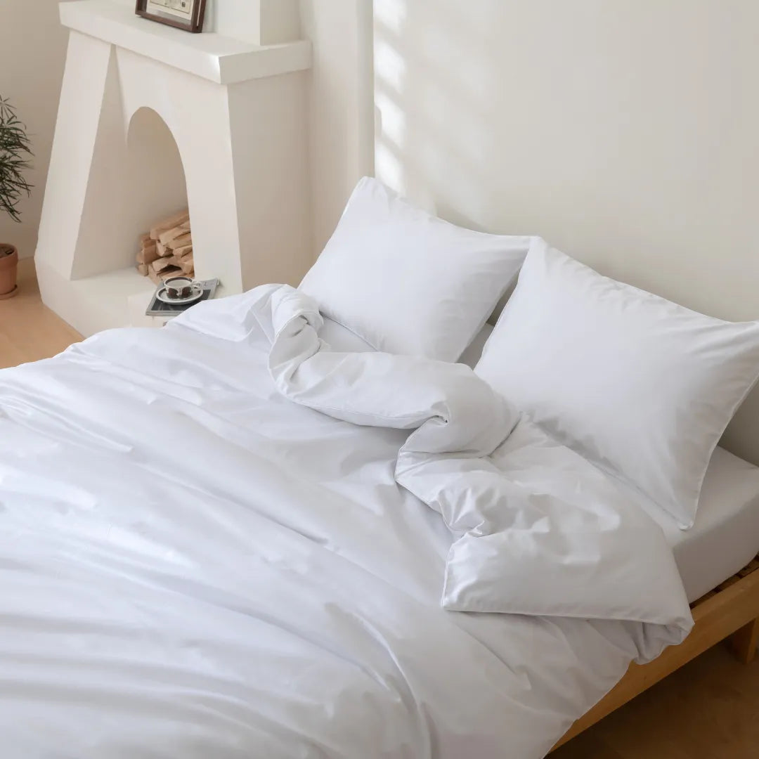 A neatly made bed with white bedding and Linenly's Luxe Cotton Sateen Pillowcase Set is placed near a white fireplace with stacked firewood. Soft daylight fills the room, creating a cozy and inviting bedroom atmosphere.