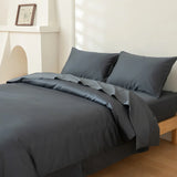 A minimalist room features a wooden bed dressed in Linenly's Luxe Cotton Sateen Quilt Cover in Charcoal, paired with a comforter and two pillows, set against white walls with a small mantel and framed picture.