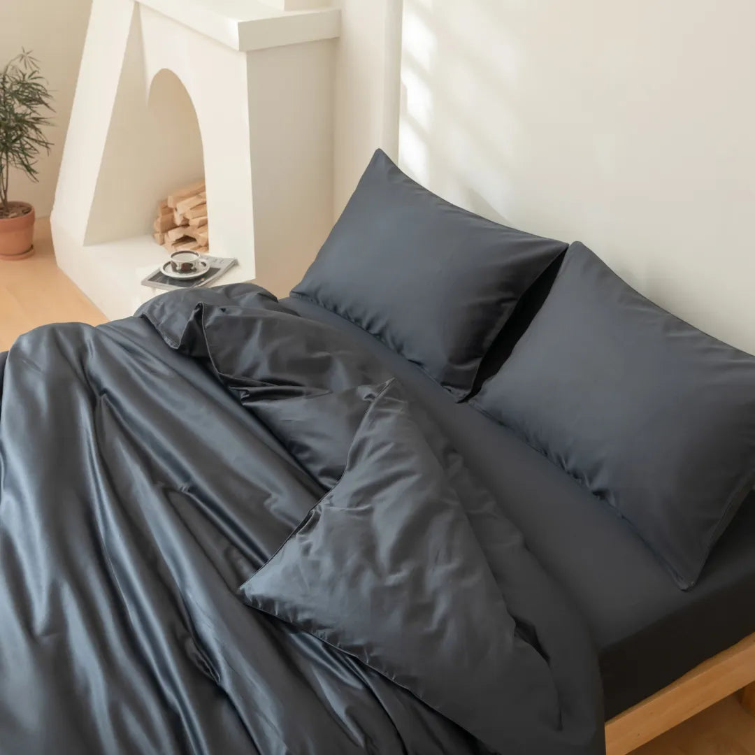 A modern bedroom features a neatly made bed with the Linenly Luxe Cotton Sateen Quilt Cover in Charcoal and dark gray pillows, positioned beside a white fireplace with stacked firewood, a small plant, and a tray holding a cup and saucer on the floor.