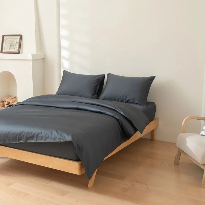 A neatly made bed with dark grey bedding, featuring a Linenly Luxe Sateen Quilt Cover in Charcoal, in a bright, minimalistic bedroom setting.