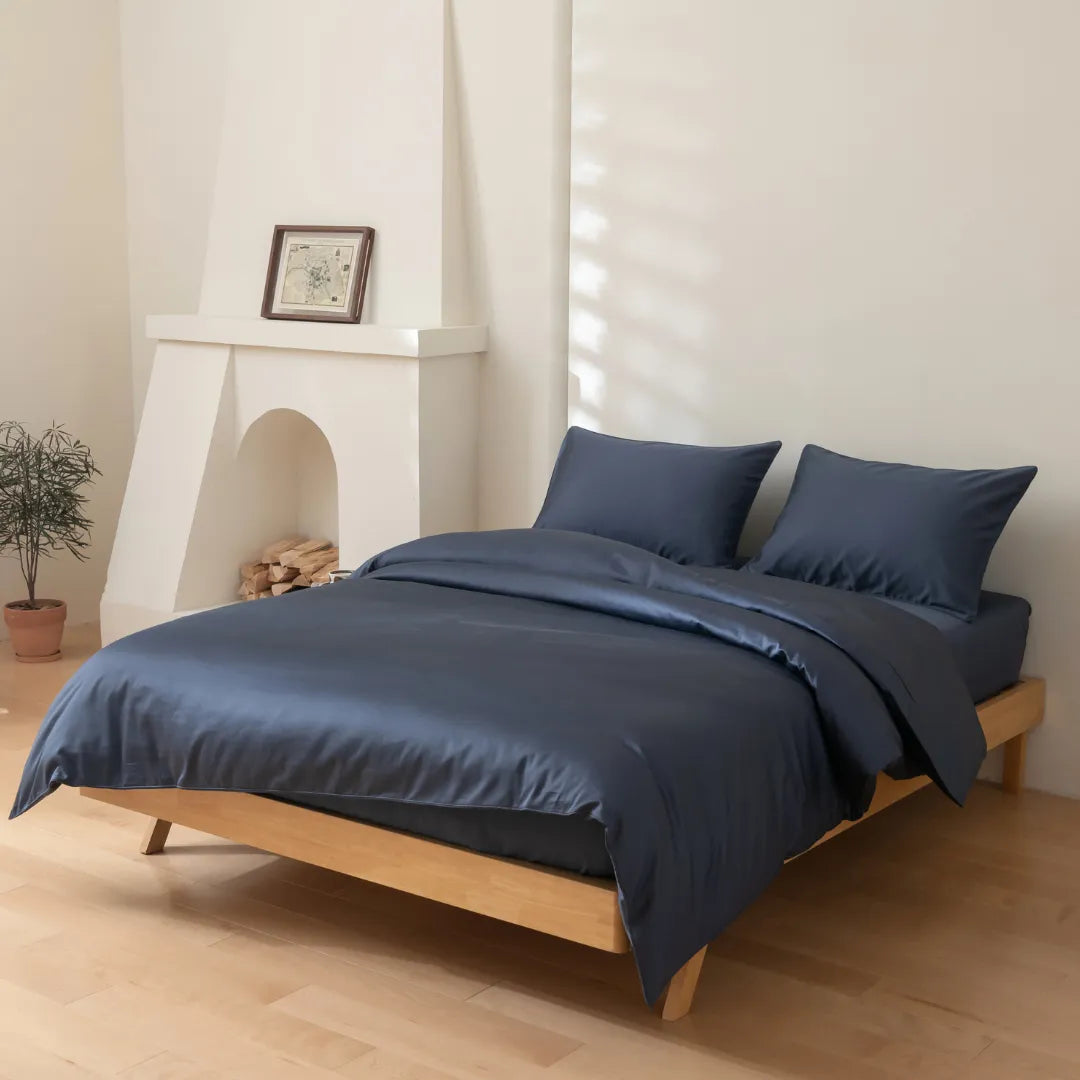 A minimalist room with light wood floors features a neatly made bed dressed in the Linenly Luxe Cotton Sateen Quilt Cover in Midnight. Two pillows, a small potted plant, and a white fireplace with framed artwork complete the décor.