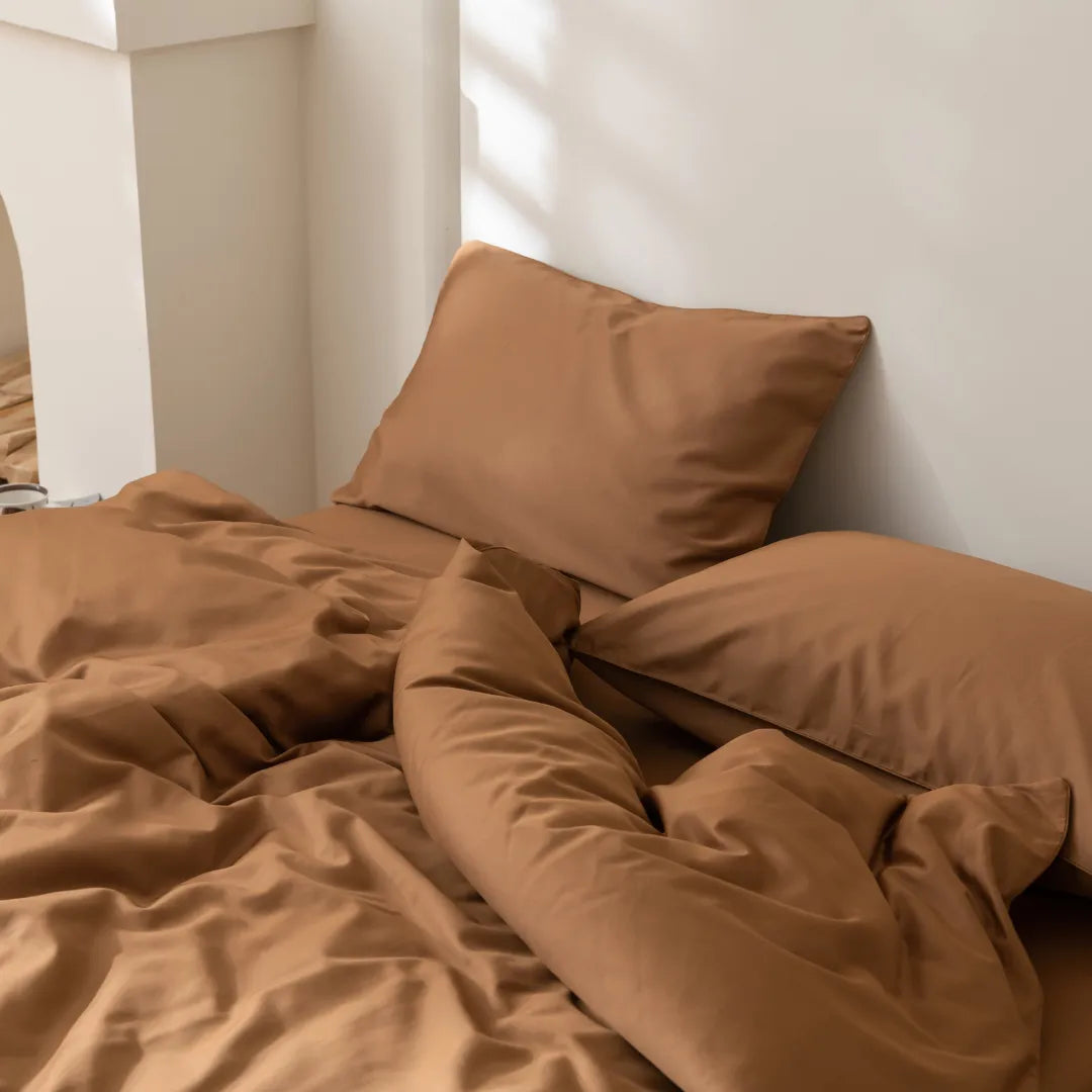 A bed with Linenly's Luxe Cotton Sateen Quilt Cover in terracotta and two pillows is set against a white wall, with sunlight highlighting the warm, inviting look of the tan bedding.