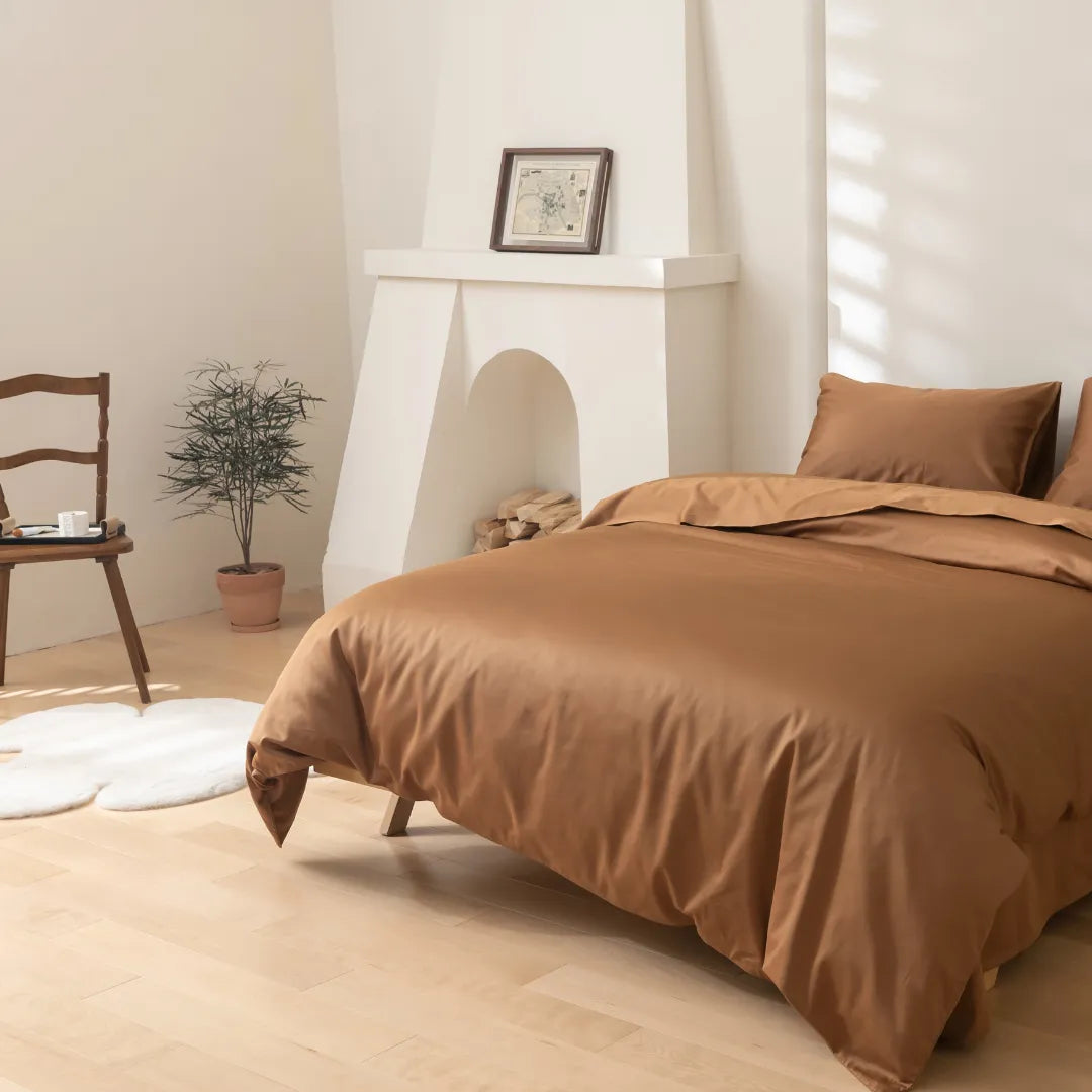 A minimalist bedroom features the Linenly Luxe Cotton Sateen Quilt Cover in terracotta, paired with brown bedding, on a neatly made bed. Light wood floors, a white fireplace, framed art, and natural light complete the serene space.