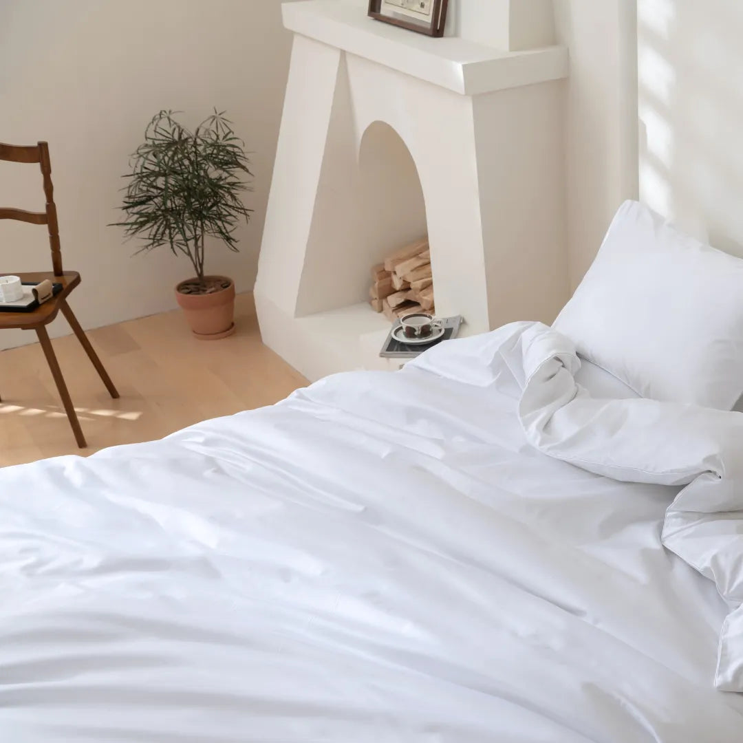 A bright room features a neatly made bed with Linenly's Luxe Cotton Sateen Quilt Cover in white and matching pillows. In the background, there’s a potted plant, wooden chair with tray, and a decorative fireplace with stacked logs.