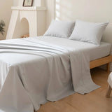 A neatly made bed features the Linenly Luxe Cotton Sateen Sheet Set in Silver with two matching pillows on a wooden frame, set in a bright, minimalist bedroom with light walls and a small plant in the corner.