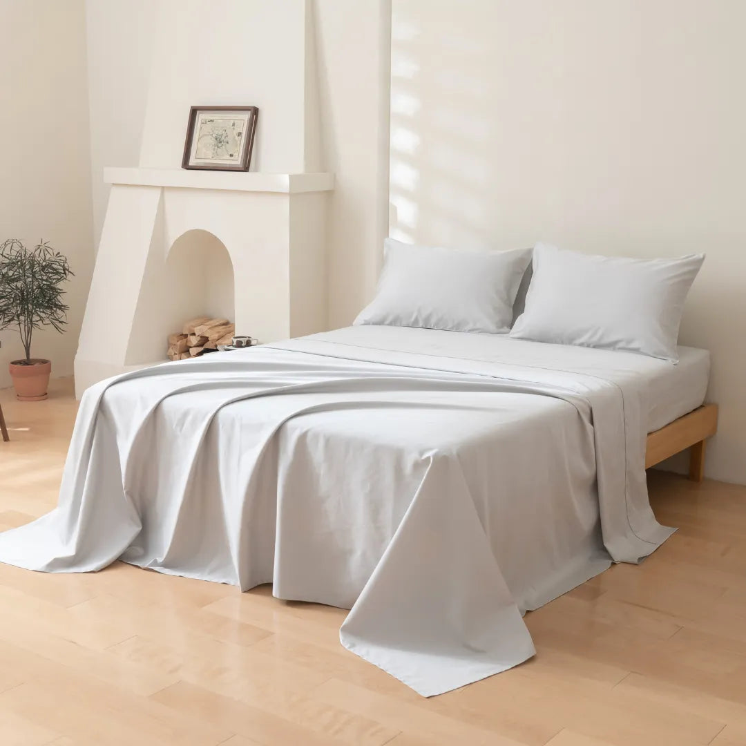 A neatly made bed with a Linenly Luxe Sateen Sheet Set in Silver, in a bright, minimalistic room with a decorative fireplace and a potted plant.