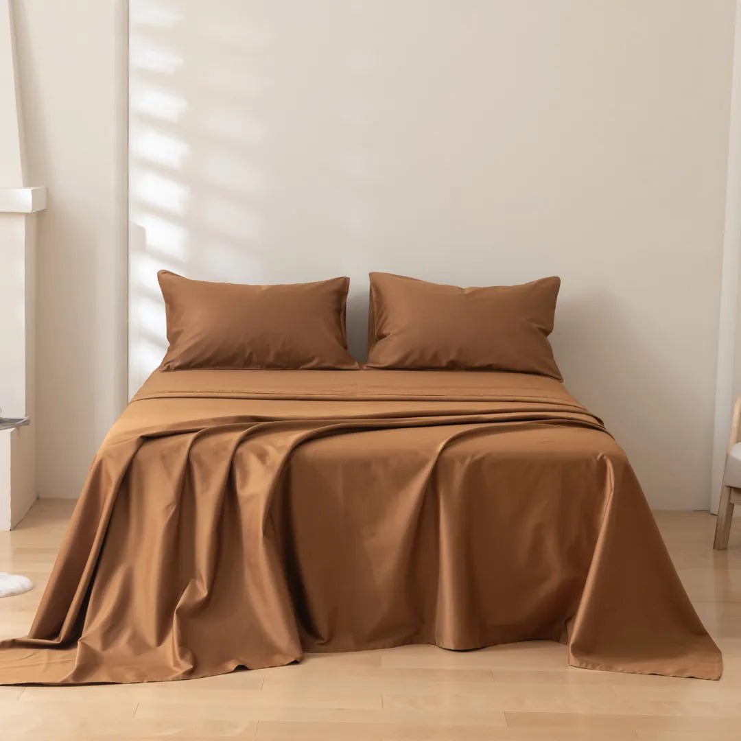 A neatly made bed features the Linenly Luxe Cotton Sateen Sheet Set in terracotta with two matching pillows, set against a light wall and wooden floor in a minimalist room where natural light casts soft shadows.