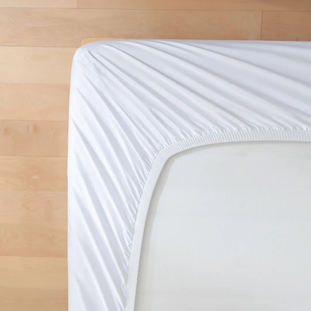 A close-up, top-down view of the Luxe Cotton Sateen Sheet Set in White by Linenly neatly covering the corner of a mattress, with a light wooden floor in the background.