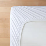 A close-up, top-down view of the Luxe Cotton Sateen Sheet Set in White by Linenly neatly covering the corner of a mattress, with a light wooden floor in the background.