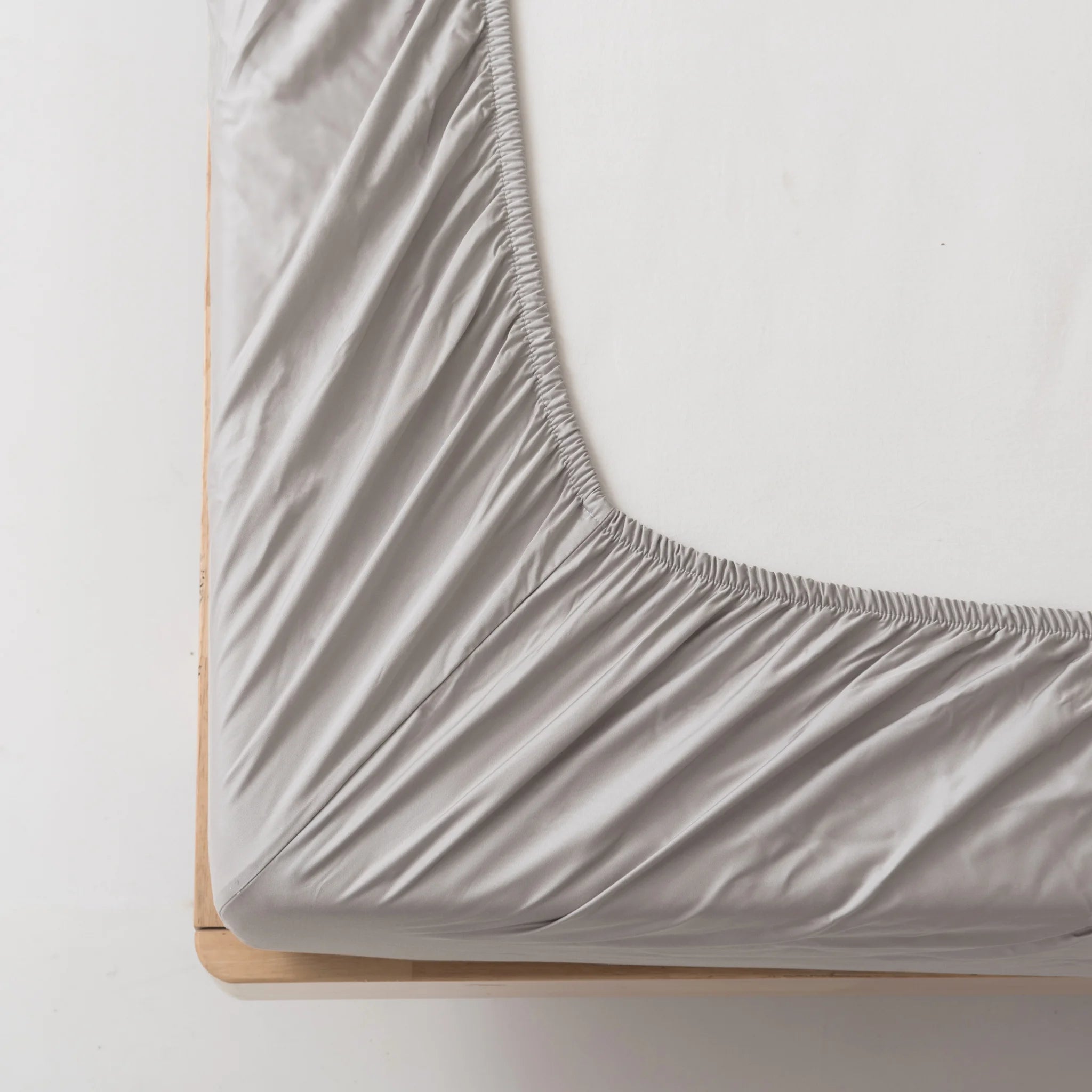 A close-up of a bed corner shows the Linenly Bamboo Fitted Sheet Set - Silver stretched over a mattress on a light wood frame. Elastic edges keep this sustainable luxury sheet securely in place.