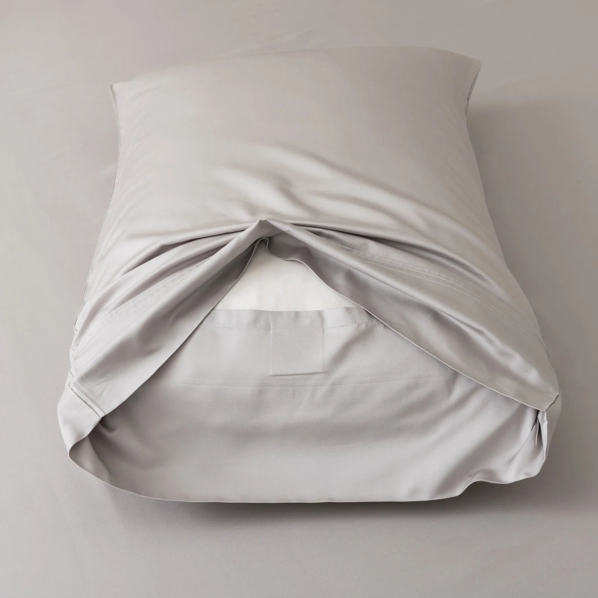 A neatly made pillow, dressed in Linenly's Bamboo Pillowcase Set - Silver, sits on a matching gray surface. The silky-smooth organic bamboo pillowcase is partially open at the back, revealing a white pillow insert inside.