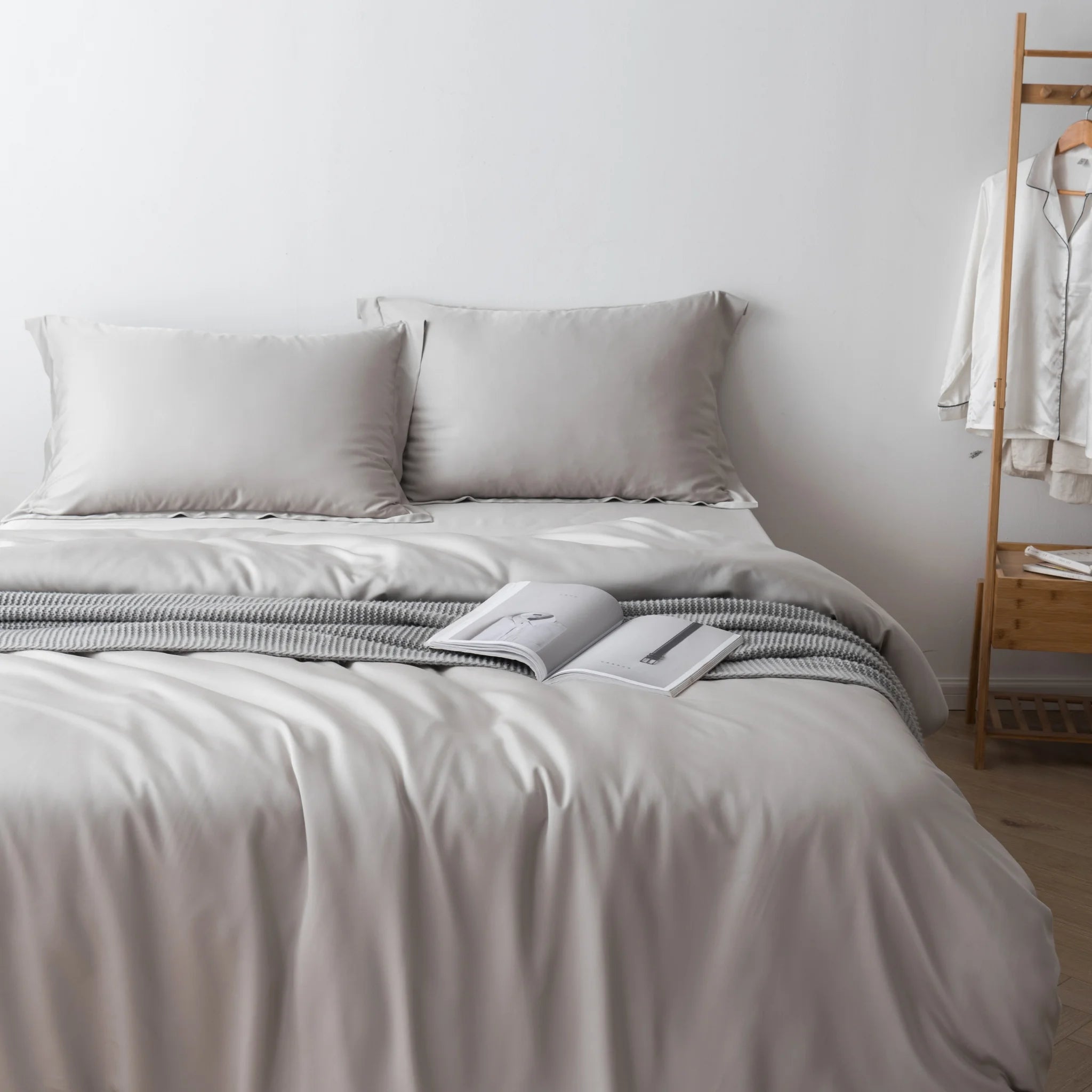 A neatly made bed features the Linenly Bamboo Quilt Cover Set in Silver with two pillows, a striped blanket at the foot, and an open book. In the background, a wooden rack displays white pajamas against a plain white wall.