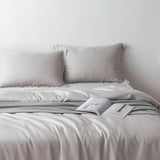A neatly made bed with the Linenly Bamboo Quilt Cover Set in Silver and two pillows is accented by a gray knitted throw and an open book, all against a plain white wall. Enjoy ultimate comfort and cooling for restful nights.