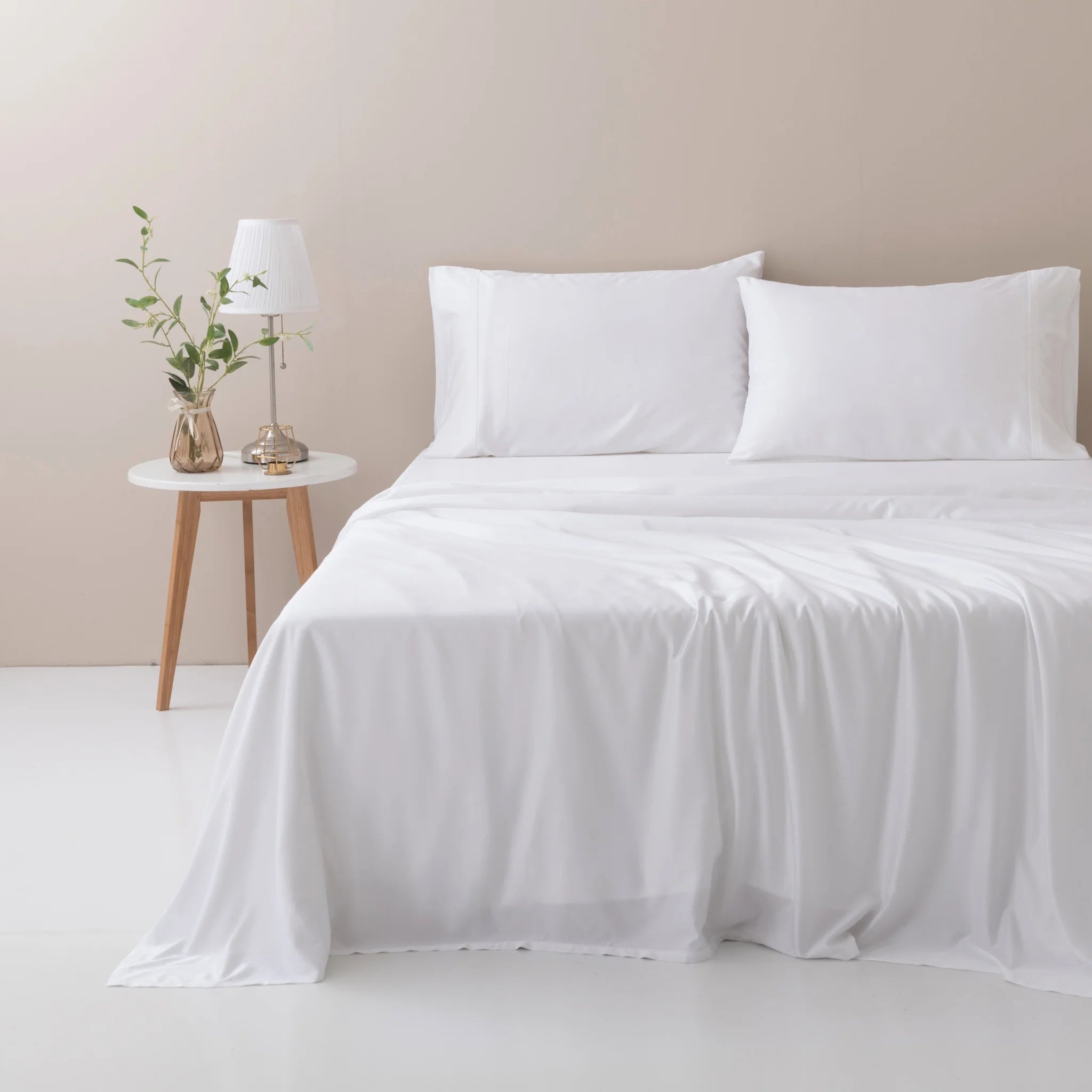 A neatly made bed with Linenly's ultra-soft Bamboo Sheet Set - White and pillows sits beside a small round table with a white lamp and green plant, all set against a beige wall in this breathable, minimalist modern bedroom.